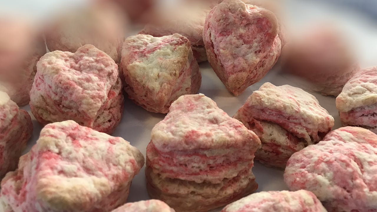 Heart-shaped biscuits with light pink swirls are arranged closely together on a tray. They have a flaky texture and a soft, golden-brown finish.