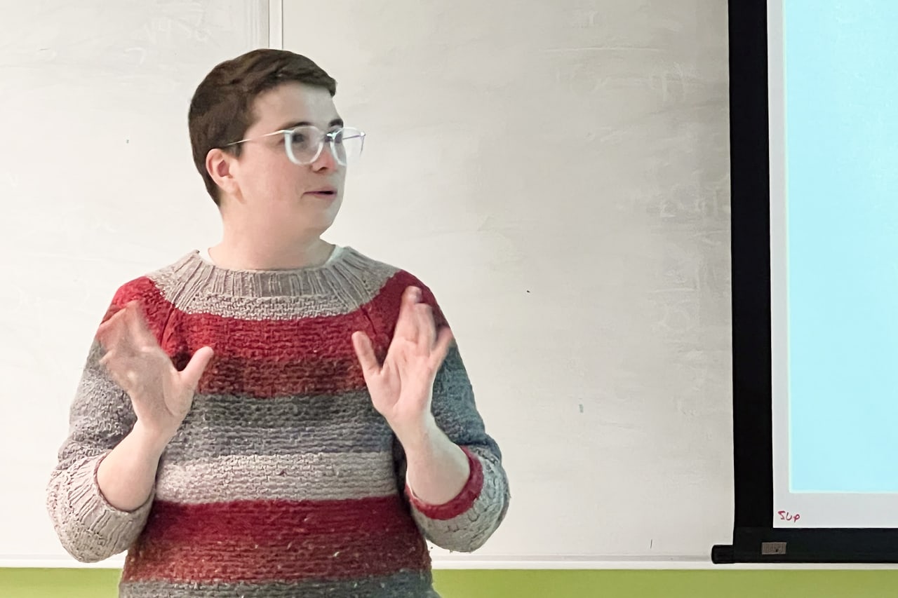 Owen Cameron (he/him) stands at the front of a classroom giving a presentation titled ‘Becoming a Designer.’